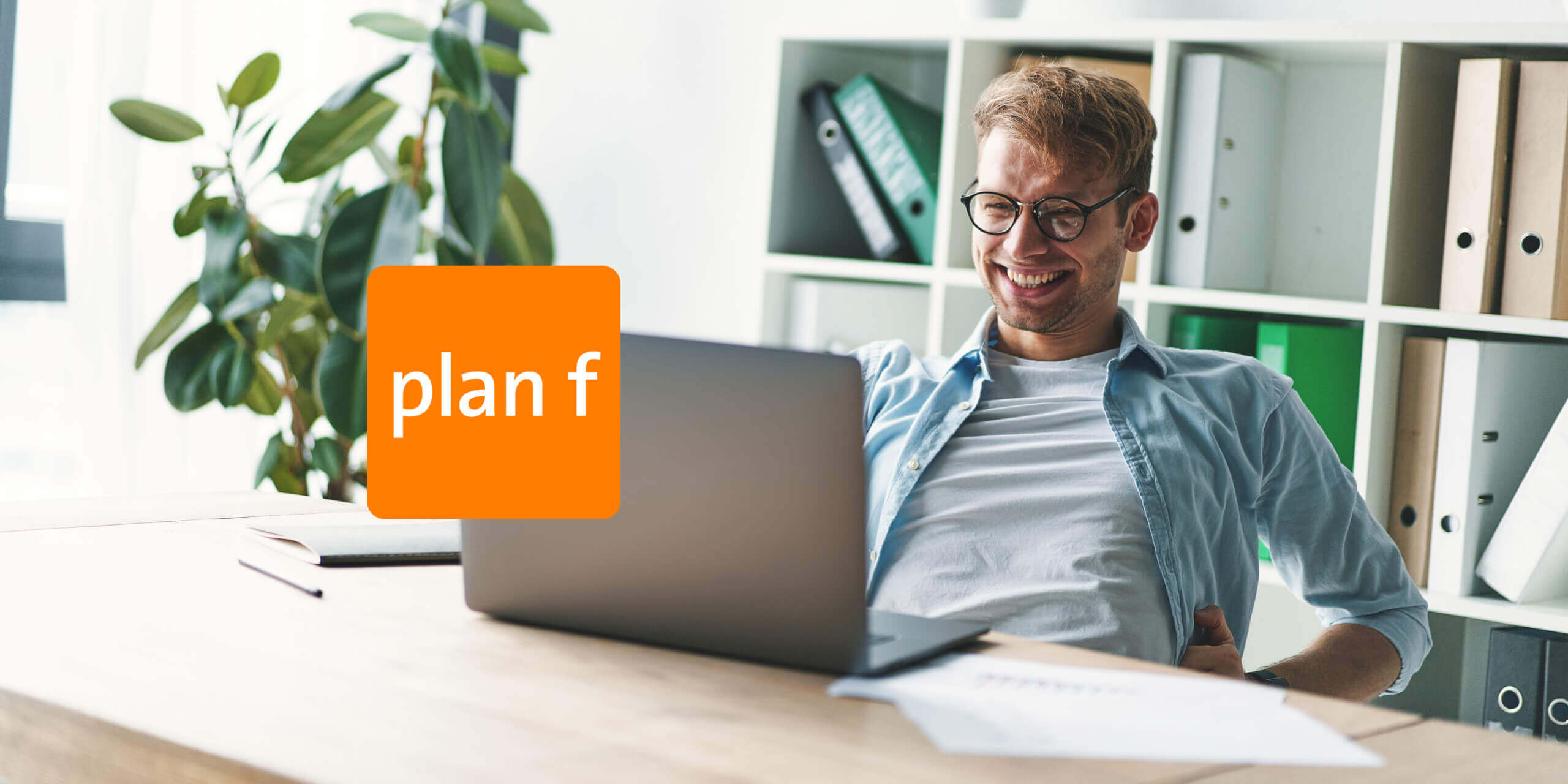 plan-f_wordpress-agentur-schweiz_3 Ein Mann mit Brille sitzt lächelnd an einem Schreibtisch und schaut auf einen Laptop in einem Büro mit dem plan f-Logo.